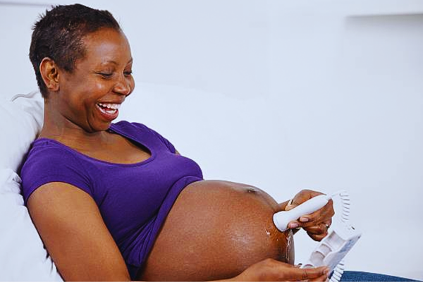 Close-up of a joyful pregnant African American woman listening to her baby with a fetal doppler, a contrast to the considerations around the Cost of Non-Invasive Prenatal Tests for early screening