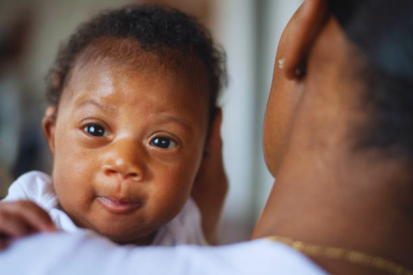 A new mother holds her infant son over her shoulder, with the baby boy wide awake, holding his head up, and showing a neutral expression, symbolizing awareness about sickle cell anemia in newborns.