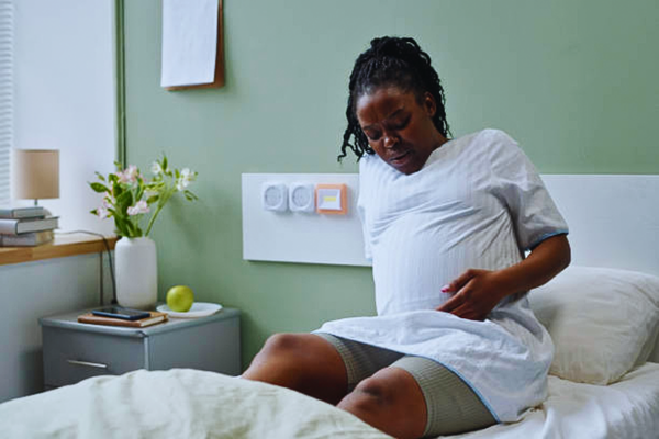 A photorealistic image of an African American pregnant woman sitting on a hospital bed, holding her belly with a look of discomfort, surrounded by flowers and medical equipment, symbolizing sickle cell pregnancy complications and the need for specialized maternal care.
