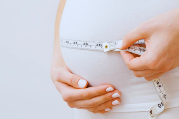Side view of a pregnant woman at 20 weeks gently measuring her belly against a gray background, illustrating body changes during pregnancy and considerations related to high BMI."