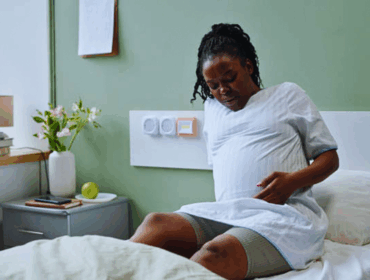 Sickle Cell Pregnancy Complications Risks, Management, and Care for Expectant Mothers A photorealistic image of an African American pregnant woman sitting on a hospital bed, holding her belly with a look of discomfort, surrounded by flowers and medical equipment, symbolizing sickle cell pregnancy complications and the need for specialized maternal care.