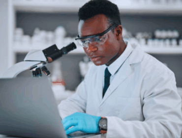 Black male scientist working with a microscope and laptop in a medical research lab, illustrating advanced laboratory procedures used in different types of DNA test analysis.