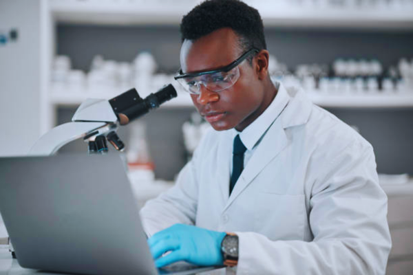 Black male scientist working with a microscope and laptop in a medical research lab, illustrating advanced laboratory procedures used in different types of DNA test analysis.